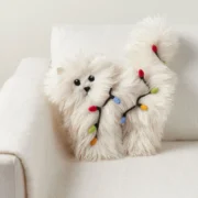 A fluffy white cat-shaped pillow wrapped in colorful Christmas lights on a white armchair.