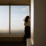 A woman standing beside sunlit windows overlooking a city.