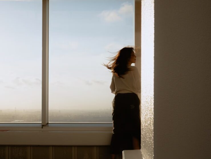 A woman standing beside sunlit windows overlooking a city.