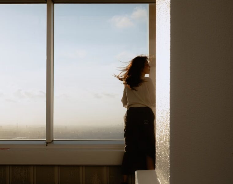 A woman standing beside sunlit windows overlooking a city.