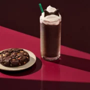 Starbucks Black Cat Frappuccino topped with whipped cream and chocolate cat ears beside a chocolate Halloween cookie.