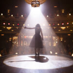 A woman standing on a theater stage beneath a bright spotlight.