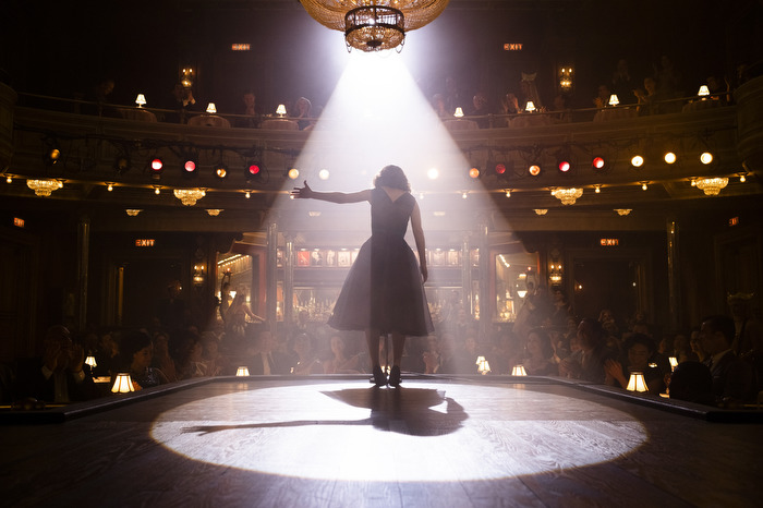 A woman standing on a theater stage beneath a bright spotlight.